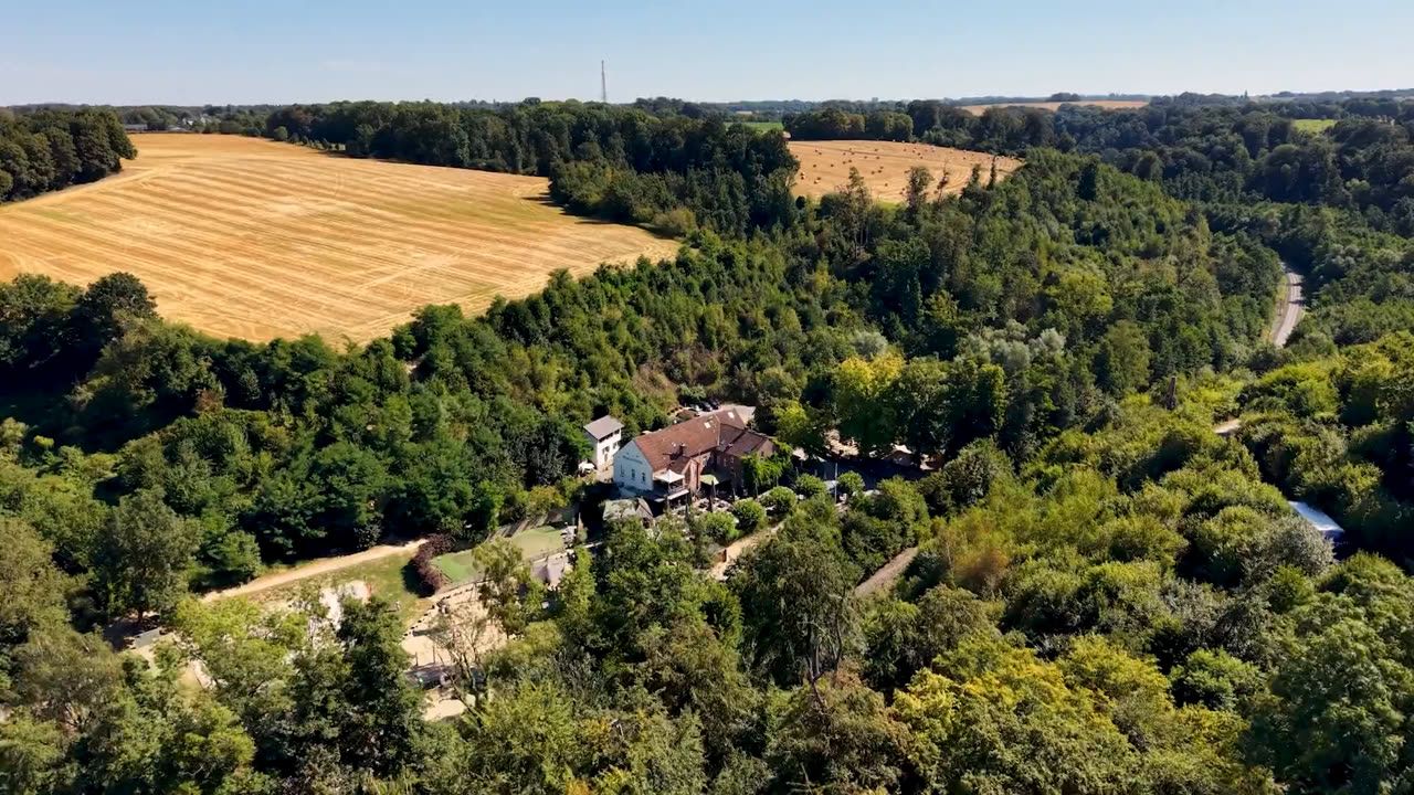 Luftbild der Auermühle in Ratingen: das Mühlengebäude im bewaldeten Angertal, dahinter abgeerntete Felder